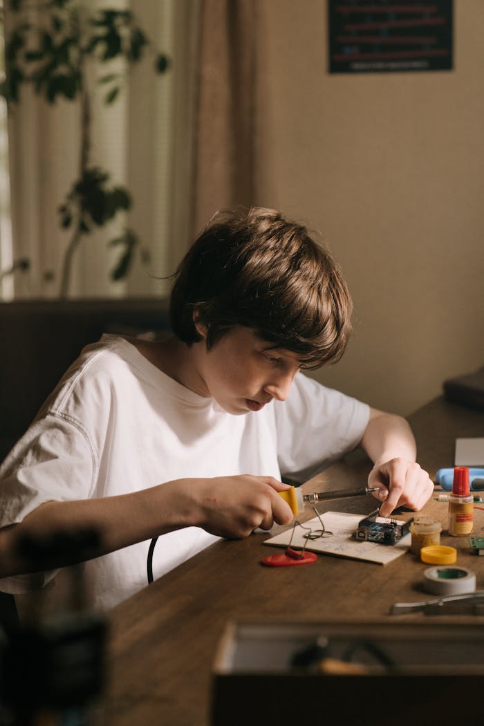 A young boy focused on soldering a printed circuit board at a desk with various electronic tools.